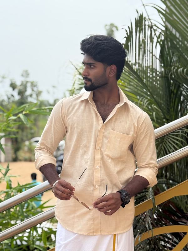 Eswar in a sandal-coloured shirt and veshti, leaning on a railing among palms